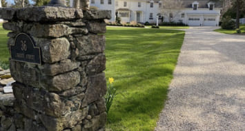 In front of a large house, a stone pillar showcases the cost-effective chipseal driveway located in Fairfield, CT.