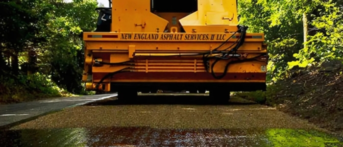 A yellow truck on a road with trees in the background, showcasing the aggregate spreading process in chip seal paving.