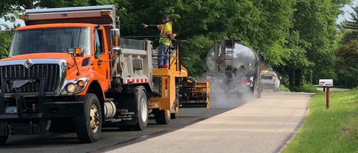 A truck drives down a rural road with a man standing on the back, illustrating chip seal paving techniques.