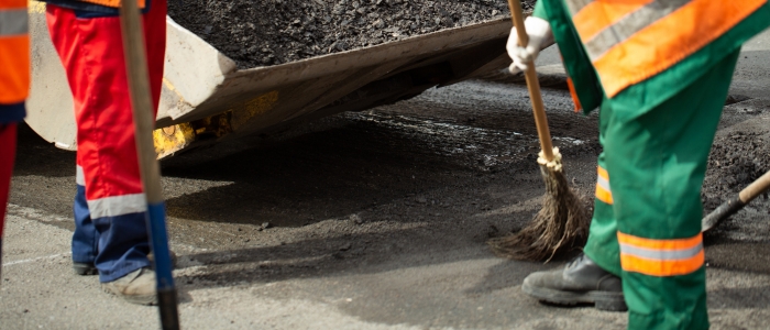 A man wearing an orange and green uniform sweeps the road, illustrating the sweeping phase of chip seal paving.