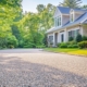 Light-colored chip seal driveway curving through a landscaped front yard toward a traditional house.