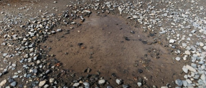 A water puddle on the ground adjacent to a road, representing maintenance needs for gravel versus chip seal.