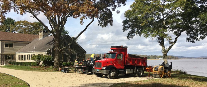A red dump truck is parked next to a tranquil lake, representing Macadam Driveways in Essex, CT.