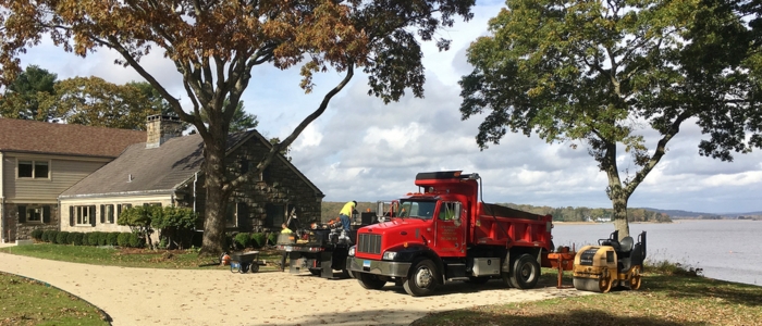 A red dump truck rests near a calm lake, framed by lush trees and scenic hills in the background.