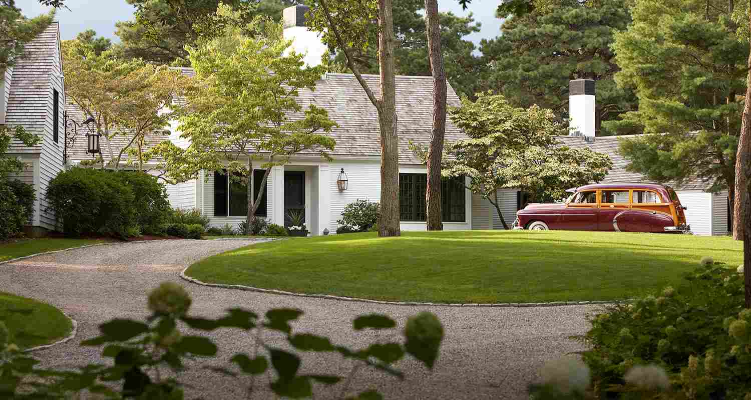 A white shingle house with a vintage woody wagon parked on a curved gravel driveway.