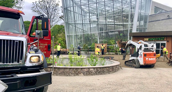 Crews using a Bobcat and red truck to pave near a large glass conservatory building.
