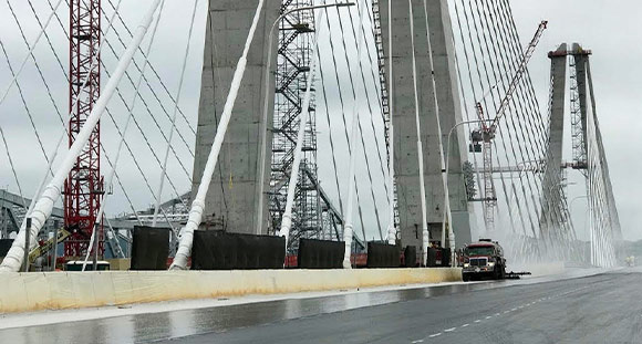 Red tanker truck spraying a dark sealant onto a bridge surface under construction.