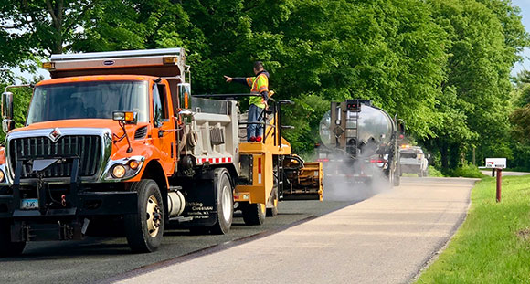 Orange dump truck and tanker truck paving a tree-lined residential road.