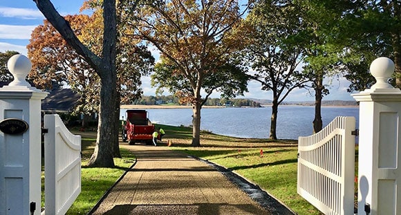 White gates open to a gravel driveway leading toward a red truck and a calm lake.